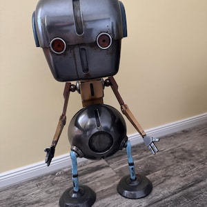 Könnte beinhalten: Handgefertigte Roboter-Skulptur mit metallisch grauem Kopf und Körper, mit roten augenähnlichen Details. Der Roboter hat bewegliche Holzarm und blaue Beine, die auf kreisförmigen Füßen stehen. Das Design hat eine Vintage-Industrie-Ästhetik.
