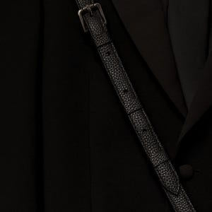 Op de afbeelding: Een zwarte blazer met een gladde stoffen afwerking en een bijpassende zwarte das, gedragen met een zwarte, getextureerde leren schouderband met een zilveren metalen gesp en twee zilveren metalen studs aan het onderste uiteinde van de band.