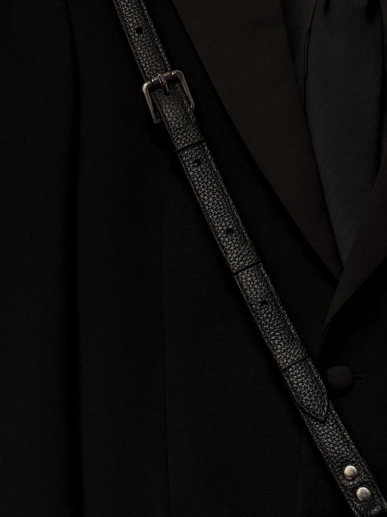 Op de afbeelding: Een zwarte blazer met een gladde stoffen afwerking en een bijpassende zwarte das, gedragen met een zwarte, getextureerde leren schouderband met een zilveren metalen gesp en twee zilveren metalen studs aan het onderste uiteinde van de band.