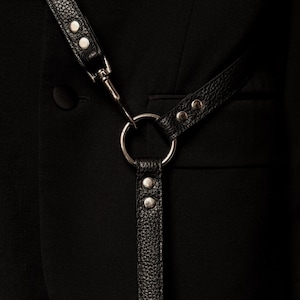 Op de afbeelding: Een zwart getextureerd lederen harnas met zilverkleurige metalen hardware, inclusief een centrale ring, klinknagels en een draaibare sluiting, gedragen over een effen zwarte blazer met een zichtbare knoop en een horizontale zakflap.