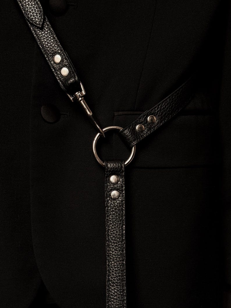 Op de afbeelding: Een zwart getextureerd lederen harnas met zilverkleurige metalen hardware, inclusief een centrale ring, klinknagels en een draaibare sluiting, gedragen over een effen zwarte blazer met een zichtbare knoop en een horizontale zakflap.