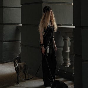 Op de afbeelding: Een zwarte jumpsuit met een harnas, een zwarte hoofddoek met patroon en een klein wit tasje aan een riem. Er zijn twee honden aanwezig, een bruine en een zwarte, beide met witte halsbanden en zwarte riemen die door de drager worden vastgehouden.