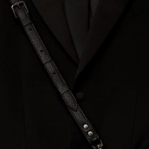 Op de afbeelding: Een zwarte blazer met een ingekeepte revers en een enkele met stof beklede knoopsluiting. Een zwarte, getextureerde leren riem met een zilveren metalen gesp en twee zilveren klinknagels loopt diagonaal over de voorkant en verbindt met een zilveren metalen ring.