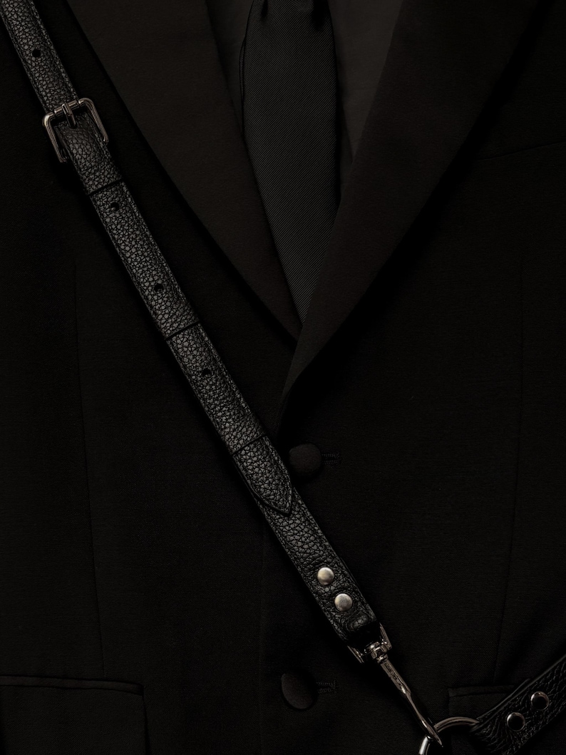 Op de afbeelding: Een zwarte blazer met een ingekeepte revers en een enkele met stof beklede knoopsluiting. Een zwarte, getextureerde leren riem met een zilveren metalen gesp en twee zilveren klinknagels loopt diagonaal over de voorkant en verbindt met een zilveren metalen ring.
