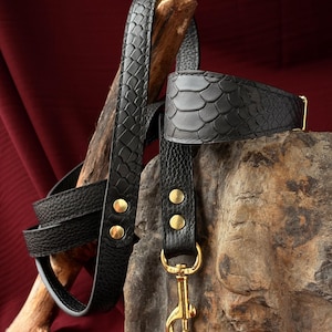 May include: A black leather dog leash and collar set featuring a textured snakeskin pattern on the outer surface. The set includes gold-toned metal hardware, including a swivel snap hook and rivets, providing a secure connection for pet walking.