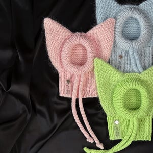 Op de afbeelding: Drie gebreide kattenoormutsen in pastelkleuren: roze, lichtblauw en groen. Elke muts heeft een cirkelvormige opening en lange trekkoorden. De mutsen worden gepresenteerd op een zwarte satijnen achtergrond, met een klein label "Made with love".