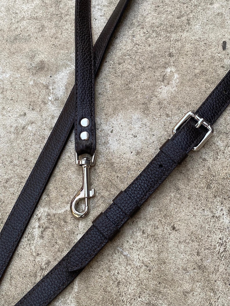Op de afbeelding: Een donkerbruine, getextureerde riem met een zilverkleurige, draaibare karabijnhaak die met twee klinknagels is bevestigd. De riem bevat een verstelbaar gespmechanisme met een schuiflus, ontworpen voor bevestiging aan tassen of accessoires.