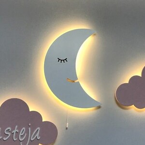 Pu&ograve; includere: Tre luci da parete in legno a forma di mezzaluna e due nuvole. La luna ha un occhio chiuso e una bocca sorridente. Una nuvola ha il nome "austeja" scritto su di essa. Tutte e tre le luci sono illuminate.