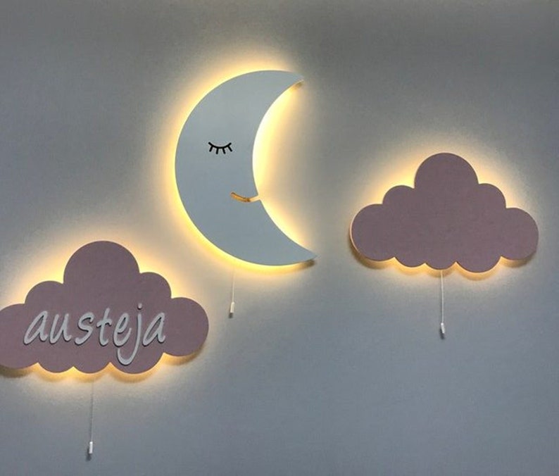 Pu&ograve; includere: Tre luci da parete in legno a forma di mezzaluna e due nuvole. La luna ha un occhio chiuso e una bocca sorridente. Una nuvola ha il nome "austeja" scritto su di essa. Tutte e tre le luci sono illuminate.