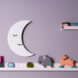 Pu&ograve; includere: Un decoro da parete a forma di mezzaluna in legno bianco con un viso sorridente e gli occhi chiusi. La luna &egrave; appesa a un muro di colore lilla chiaro sopra uno scaffale bianco. Lo scaffale ha un elefante di legno e una statuetta di cervo di legno, oltre a due blocchi rosa e bianchi che indicano "12 anni".