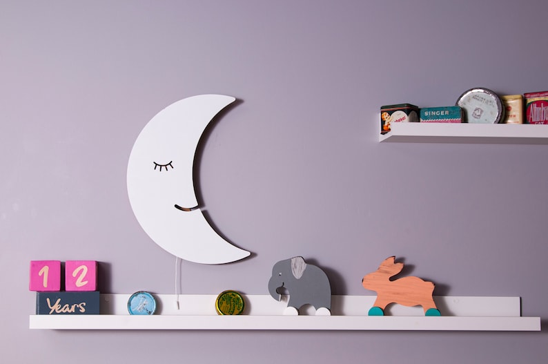 Pu&ograve; includere: Un decoro da parete a forma di mezzaluna in legno bianco con un viso sorridente e gli occhi chiusi. La luna &egrave; appesa a un muro di colore lilla chiaro sopra uno scaffale bianco. Lo scaffale ha un elefante di legno e una statuetta di cervo di legno, oltre a due blocchi rosa e bianchi che indicano "12 anni".