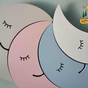 Pu&ograve; includere: Un set di quattro forme di luna crescente in legno di colore grigio, rosa, blu e bianco. Ogni luna ha un viso sorridente nero con gli occhi chiusi. Il testo "happy woody" &egrave; nell'angolo in alto a destra dell'immagine.