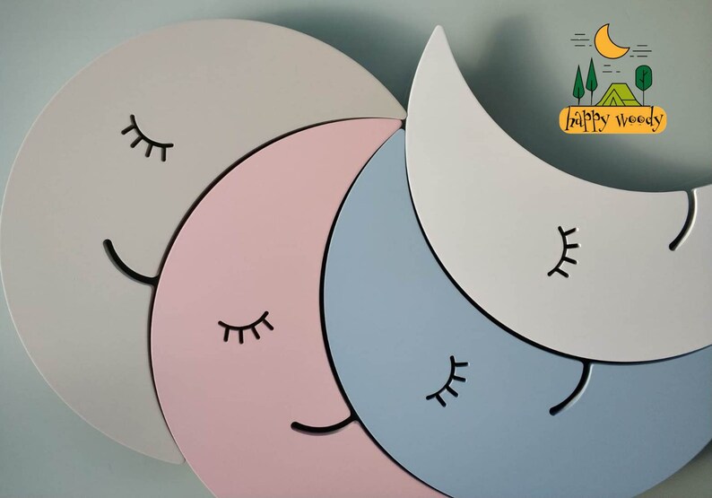 Pu&ograve; includere: Un set di quattro forme di luna crescente in legno di colore grigio, rosa, blu e bianco. Ogni luna ha un viso sorridente nero con gli occhi chiusi. Il testo "happy woody" &egrave; nell'angolo in alto a destra dell'immagine.