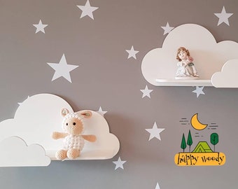 May include: Two white cloud-shaped shelves mounted on a gray wall. The top shelf has a porcelain figurine of a girl holding a flower. The bottom shelf has a crocheted white lamb toy. The wall is decorated with white stars and a green, yellow, and black drawing of a moon, trees, and a tent with the text "Happy Woody".