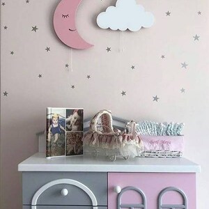 Pu&ograve; includere: Un decoro da parete a forma di mezzaluna di legno rosa con un viso sorridente e gli occhi chiusi. Un decoro da parete a forma di nuvola di legno bianco con una corda appesa &egrave; accanto alla luna. Entrambi sono su una parete rosa con adesivi a forma di stella argentata. Una cassettiera grigia e rosa con due cassetti &egrave; in primo piano. Un album fotografico e un cestino di vimini bianco con il testo "Bebe Maria" sono sopra la cassettiera.