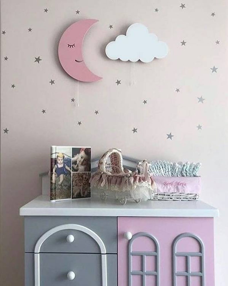 Pu&ograve; includere: Un decoro da parete a forma di mezzaluna di legno rosa con un viso sorridente e gli occhi chiusi. Un decoro da parete a forma di nuvola di legno bianco con una corda appesa &egrave; accanto alla luna. Entrambi sono su una parete rosa con adesivi a forma di stella argentata. Una cassettiera grigia e rosa con due cassetti &egrave; in primo piano. Un album fotografico e un cestino di vimini bianco con il testo "Bebe Maria" sono sopra la cassettiera.