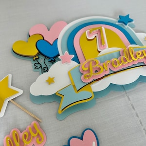 May include: A vibrant cake topper for a birthday celebration, featuring a rainbow, stars, and the number 7. The name "Bradley" is displayed in gold lettering. The design incorporates hearts and balloons in shades of blue, pink, yellow, and white.
