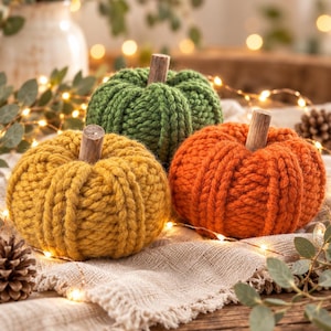 May include: Three handcrafted pumpkins made of chunky knit yarn in mustard yellow, forest green, and burnt orange. Each pumpkin features a thick, ribbed texture and a smooth, cylindrical wooden stem centered at the top of the round form.