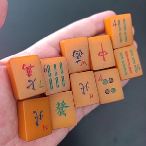 Könnte beinhalten: Eine Sammlung von zehn rechteckigen Mahjong-Spielsteinen mit bernsteinfarbener Oberfläche. Jeder Spielstein zeigt eingravierte Symbole, Schriftzeichen und Zahlen in roter, grüner, blauer und schwarzer Tinte, die verschiedene Farben und Richtungsmarkierungen darstellen.
