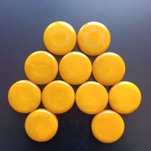 May include: A collection of ten circular, bright yellow buttons arranged in a pyramid-like formation. Each button features a smooth, glossy finish with a slightly domed surface, creating a uniform appearance across the entire set of round fasteners.