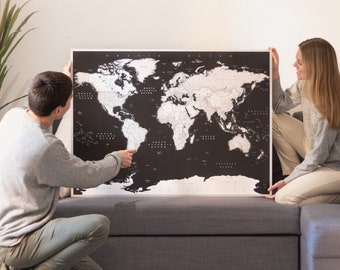 May include: Two people are hanging a large black and white world map on a wall. The map is framed and has a silver border. The map is a scratch-off map, where you can scratch off the countries you have visited.