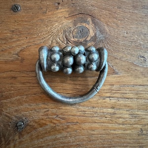 Può includere: Primo piano di una maniglia per cassetto in metallo argentato con maniglia ad anello e design decorativo a perline. L'hardware è montato su una superficie in legno con venature visibili e teste di viti, che suggeriscono un'estetica vintage o antica.