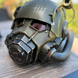 May include: An olive green tactical helmet with a weathered, distressed finish featuring rust-like details and silver paint chips. The helmet includes a black front respirator, a side-mounted circular light, and five white tally marks on the top.