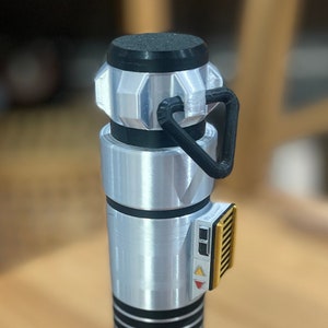May include: A silver lightsaber prop with black and gold accents. The lightsaber has a black handle with a silver blade and a gold button on the side.