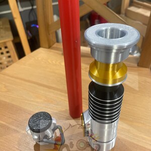 May include: A silver and gold lightsaber with a black stripe, a red lightsaber blade, and a silver cap. The lightsaber is on a wooden surface.