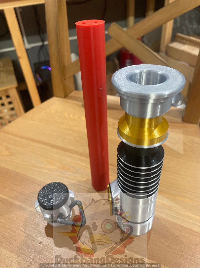 May include: A silver and gold lightsaber with a black stripe, a red lightsaber blade, and a silver cap. The lightsaber is on a wooden surface.