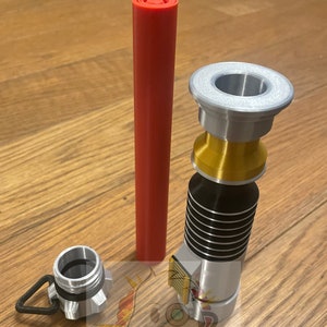 May include: A silver and gold lightsaber hilt with a black and silver grip. The hilt has a red lightsaber blade, a silver cap, and a gold ring. The hilt is on a wooden surface.