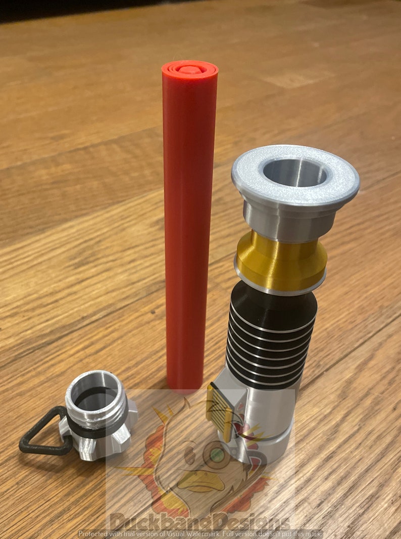 May include: A silver and gold lightsaber hilt with a black and silver grip. The hilt has a red lightsaber blade, a silver cap, and a gold ring. The hilt is on a wooden surface.