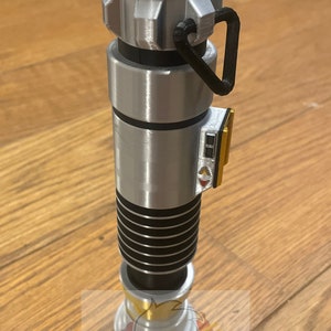 May include: A silver and black lightsaber with a black handle and a gold accent. The lightsaber has a black and silver grip and a black and silver blade.