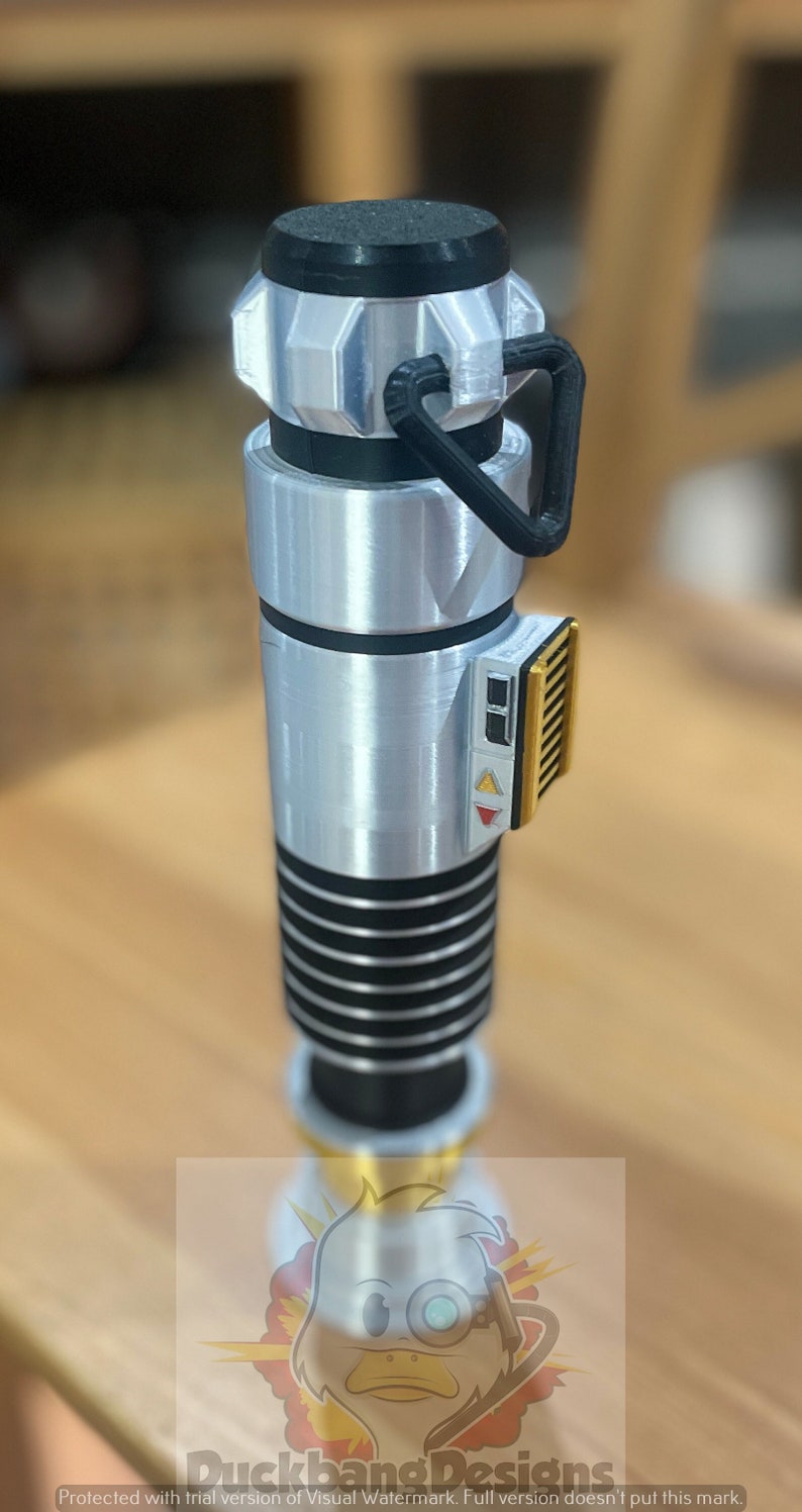 May include: A silver lightsaber prop with black and gold accents. The lightsaber has a black handle with a silver blade and a gold button on the side.