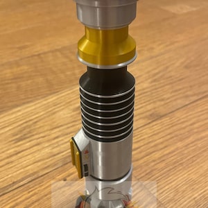 May include: A silver and gold lightsaber with black accents. The lightsaber has a red button on the top and a yellow and black switch on the side.