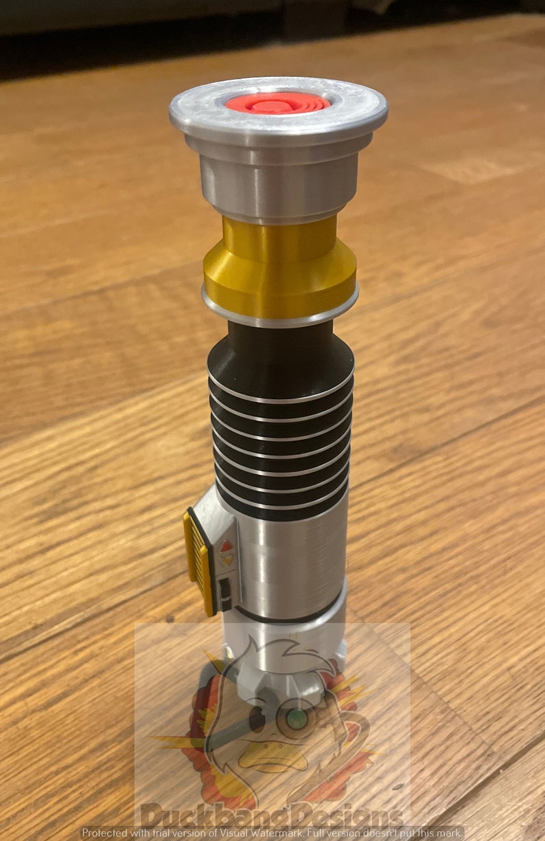 May include: A silver and gold lightsaber with black accents. The lightsaber has a red button on the top and a yellow and black switch on the side.