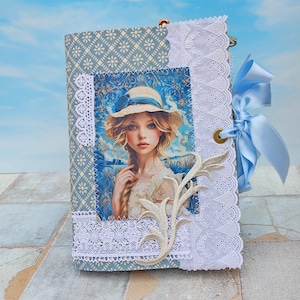 Puede incluir: Un diario hecho a mano con una cubierta de patrón floral azul y blanco. El diario presenta un retrato de una joven con sombrero, enmarcado con encaje blanco y una aplicación decorativa. Una cinta de raso azul claro está atada en el lateral.