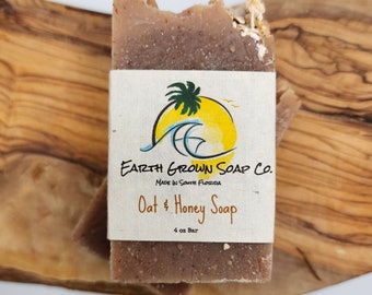 May include: A bar of Oat & Honey Soap from Earth Grown Soap Co. is shown, with a label featuring a palm tree and ocean waves. The soap is a light brown color and weighs 4 oz. The soap is on a wooden surface.