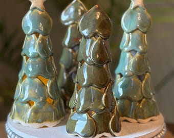 May include: Four ceramic tree-shaped sculptures with a glossy, multi-toned green glaze. Each tree features layered, heart-shaped leaves and is topped with a small, white star. The structures are hollow, allowing light to shine through the gaps.