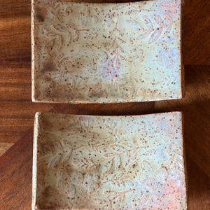 Może przedstawiać: Dwie prostokątne ceramiczne miseczki z jasnobeżowym szkliwem i nakrapianym wykończeniem. Powierzchnia każdej miseczki posiada tłoczony botaniczny wz&oacute;r liści i łodyg w ciemniejszym, czerwonobrązowym odcieniu o wyraźnej fakturze.