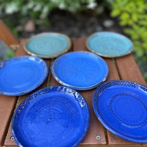 May include: A collection of handmade ceramic plates in shades of blue and turquoise. The plates have a textured surface and a rustic, artisanal look. Some plates have a decorative rim.