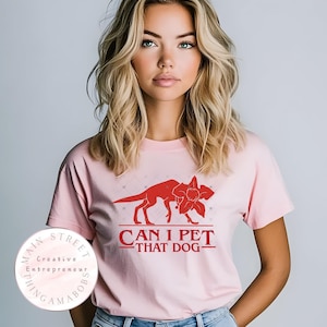 May include: A light pink t-shirt featuring a red graphic of a dog-like creature and a flower, with the text "CAN I PET THAT DOG-". The shirt is a crew neck style.