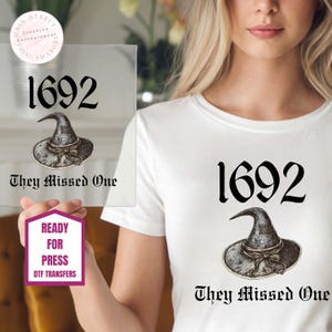 May include: A white t-shirt featuring a graphic print with the number 1692 in a black serif font, a detailed illustration of a grey witch hat, and the text They Missed One in a black gothic-style font centered below the hat illustration.