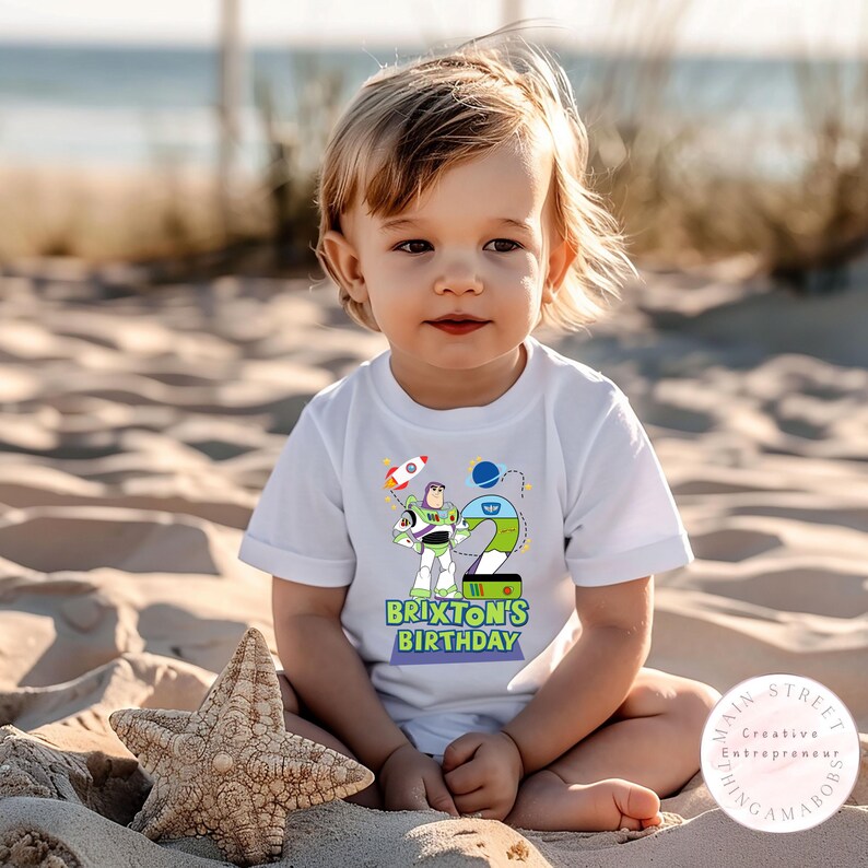 K&ouml;nnte beinhalten: Ein wei&szlig;es T-Shirt mit einer bunten Grafik von Buzz Lightyear, einer Rakete und der Zahl 2. Der Text "Brixon's Birthday" ist darunter gedruckt. Ein Seestern ist im Vordergrund. Das Kind sitzt im Sand.