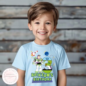 K&ouml;nnte beinhalten: Hellblaues T-Shirt mit einer Grafik von Buzz Lightyear, einer Rakete und der Zahl 3. Der Text "BRIXTON'S BIRTHDAY" ist darunter abgebildet. Das T-Shirt wird von einem kleinen Kind getragen.