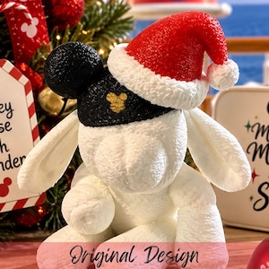 May include: A white plush animal toy wearing a black cap with round ears and a red Santa hat with white trim. A tag reads Disney Cruise Fish Extender, and a white sign features the text Making Magic at Sea with a black Mickey Mouse silhouette.