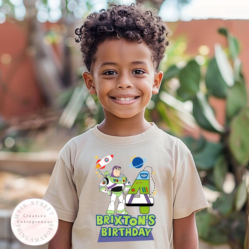 K&ouml;nnte beinhalten: Beigefarbenes Kurzarm-T-Shirt mit einem bunten Aufdruck eines Cartoon-Astronauten, einer Rakete und dem Text "Brixon's Birthday". Das T-Shirt wird von einem jungen Kind getragen.