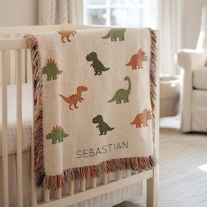 May include: A beige baby blanket with various dinosaur illustrations in shades of green, orange, and brown. The blanket is draped over a white crib and has a fringed edge. The name "SEBASTIAN" is printed on the blanket.