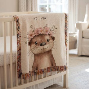 Könnte beinhalten: Eine beige Babydecke mit farbenfrohem Fransen, die über einem weißen Kinderbett drapiert ist. Die Decke zeigt eine Aquarellillustration eines Otters mit einem Blumenkranz. Der Name "OLIVIA" ist über dem Otter aufgedruckt.