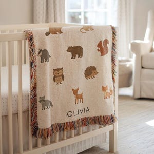 May include: A beige baby blanket with woodland animal illustrations, including a bear, squirrel, and fox. The blanket has a fringed edge with brown, orange, and red threads. The name "OLIVIA" is printed in black letters on the blanket.